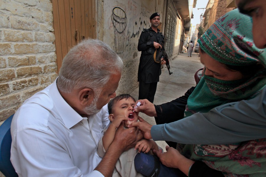 A police officer stands guard as a health worker, right, administers a polio vaccine to a child in a neighbourhood of Peshawar, Pakistan, Monday, Oct. 28, 2024. (AP Photo/Mohammad Sajjad)