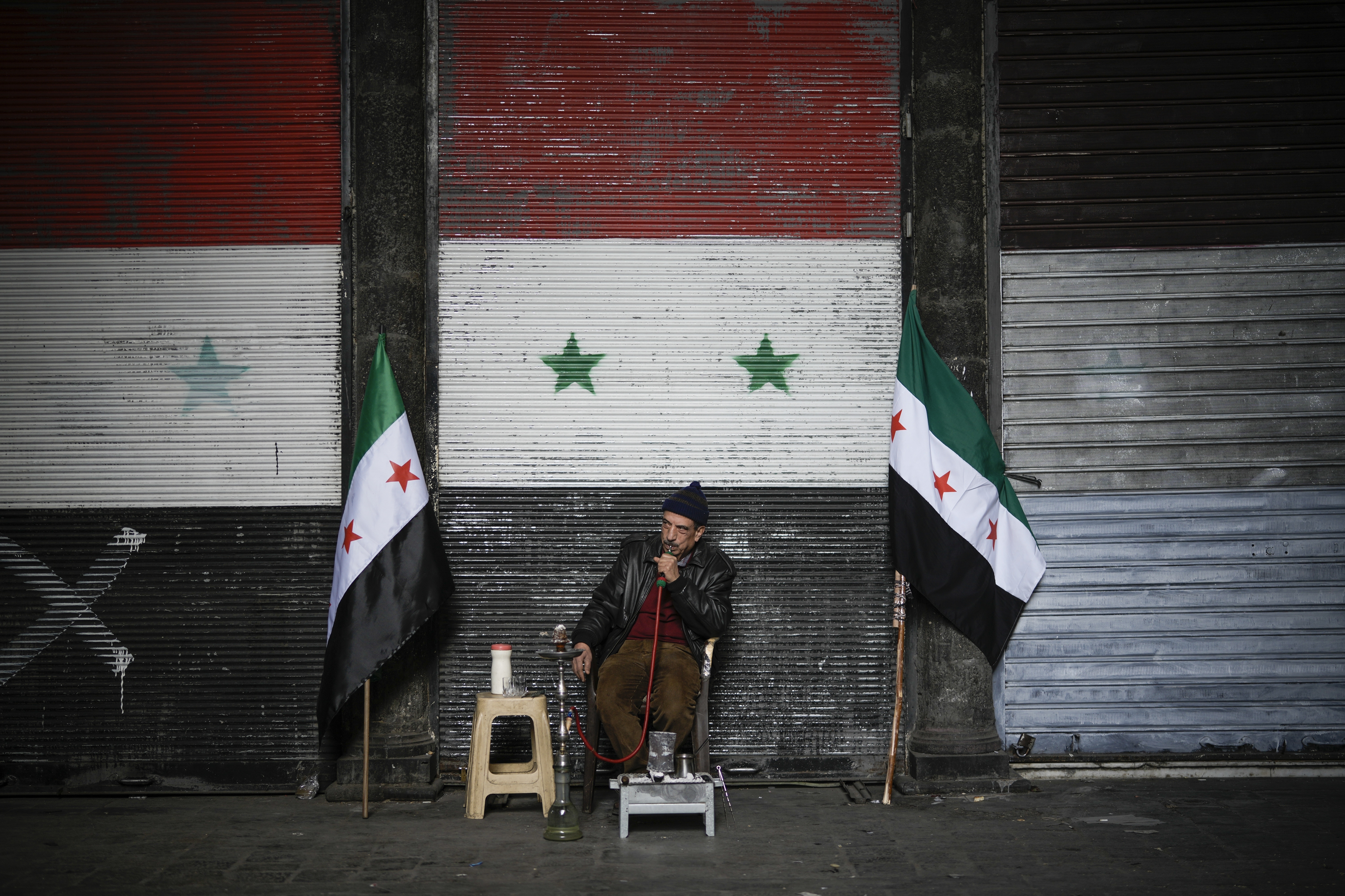 A man smokes a water pipe next to two Syrian "revolutionary" flags in front of a door still adorned with the official flag of the ousted government, near the Umayyad Mosque ahead of Friday prayers in Damascus, Syria, Friday, Dec. 20, 2024. (AP Photo/Leo Correa)