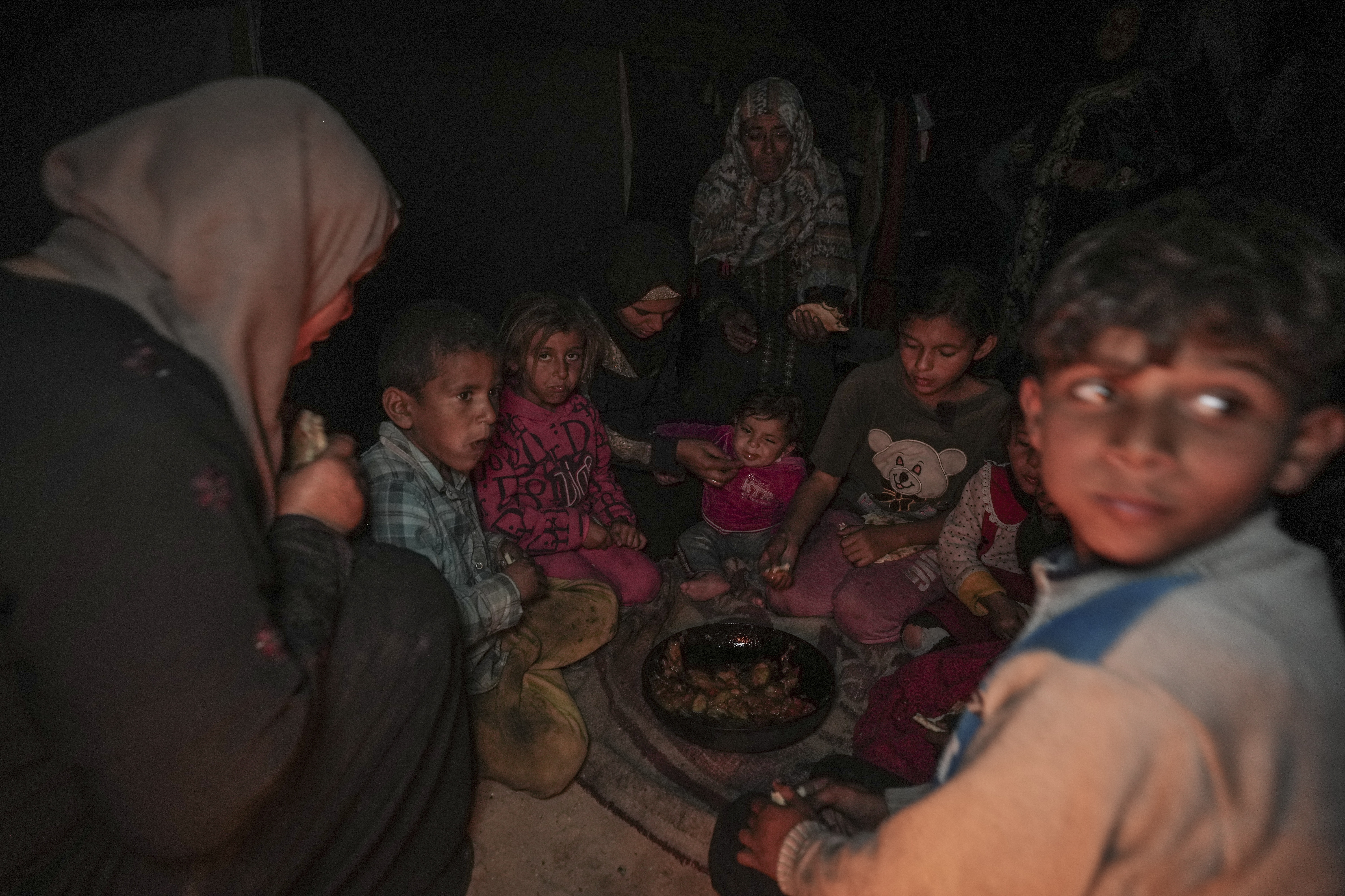 Amani Abu Zarada, fourth from left, feeds one of her children with fried zucchini made over a fire made of paper and cardboard scraps outside their tent in a camp in Khan Younis, Gaza Strip, Thursday, Dec. 19, 2024. (AP Photo/Abdel Kareem Hana)