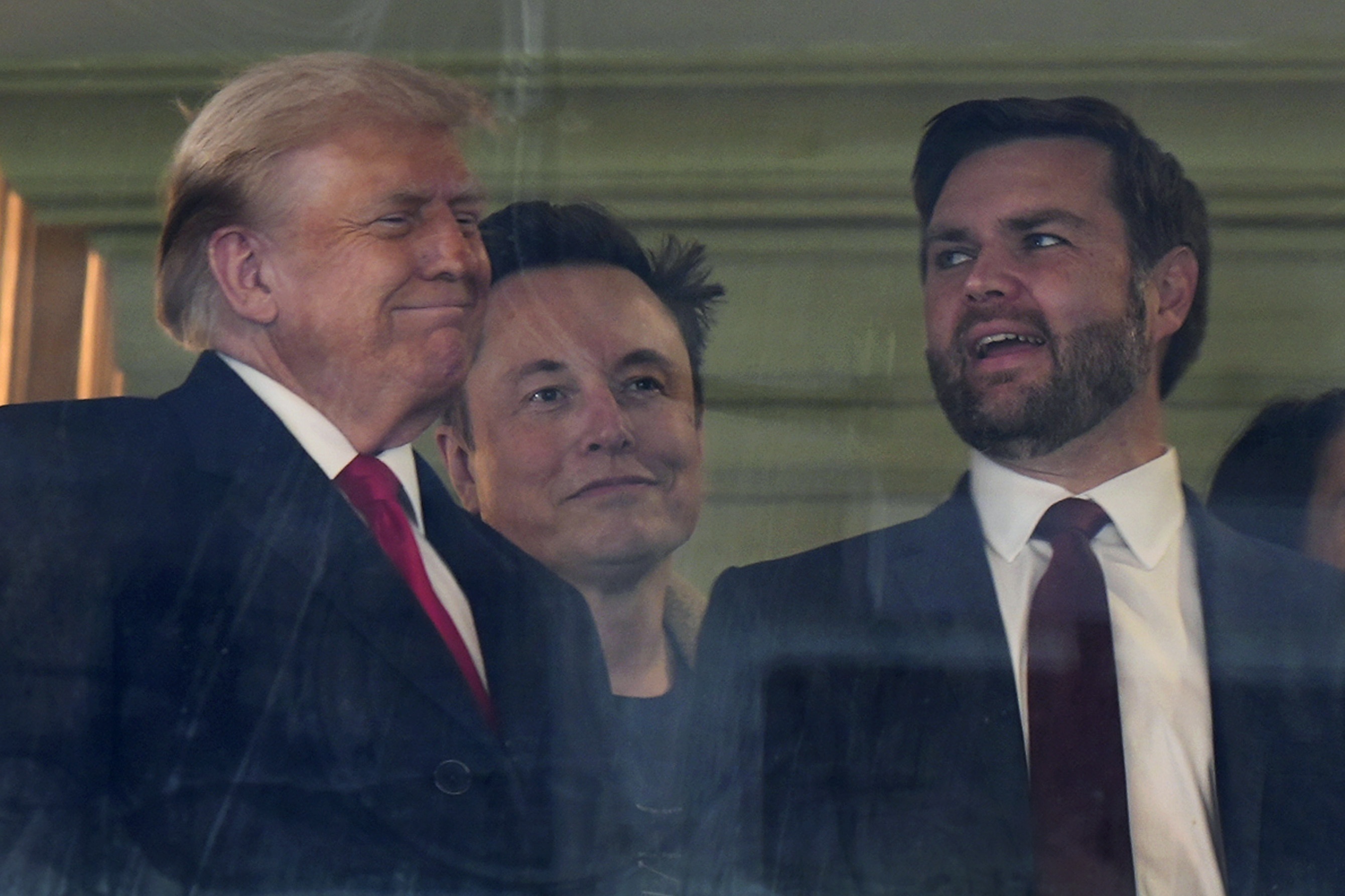 In this picture taken through a window, President-elect Donald Trump, from left, Trump's pick for the planned Department of Government Efficiency Elon Musk and Vice President-elect JD Vance attend the NCAA college football game between Army and Navy at Northwest Stadium in Landover, Md., Saturday, Dec. 14, 2024. (AP Photo/Stephanie Scarbrough)