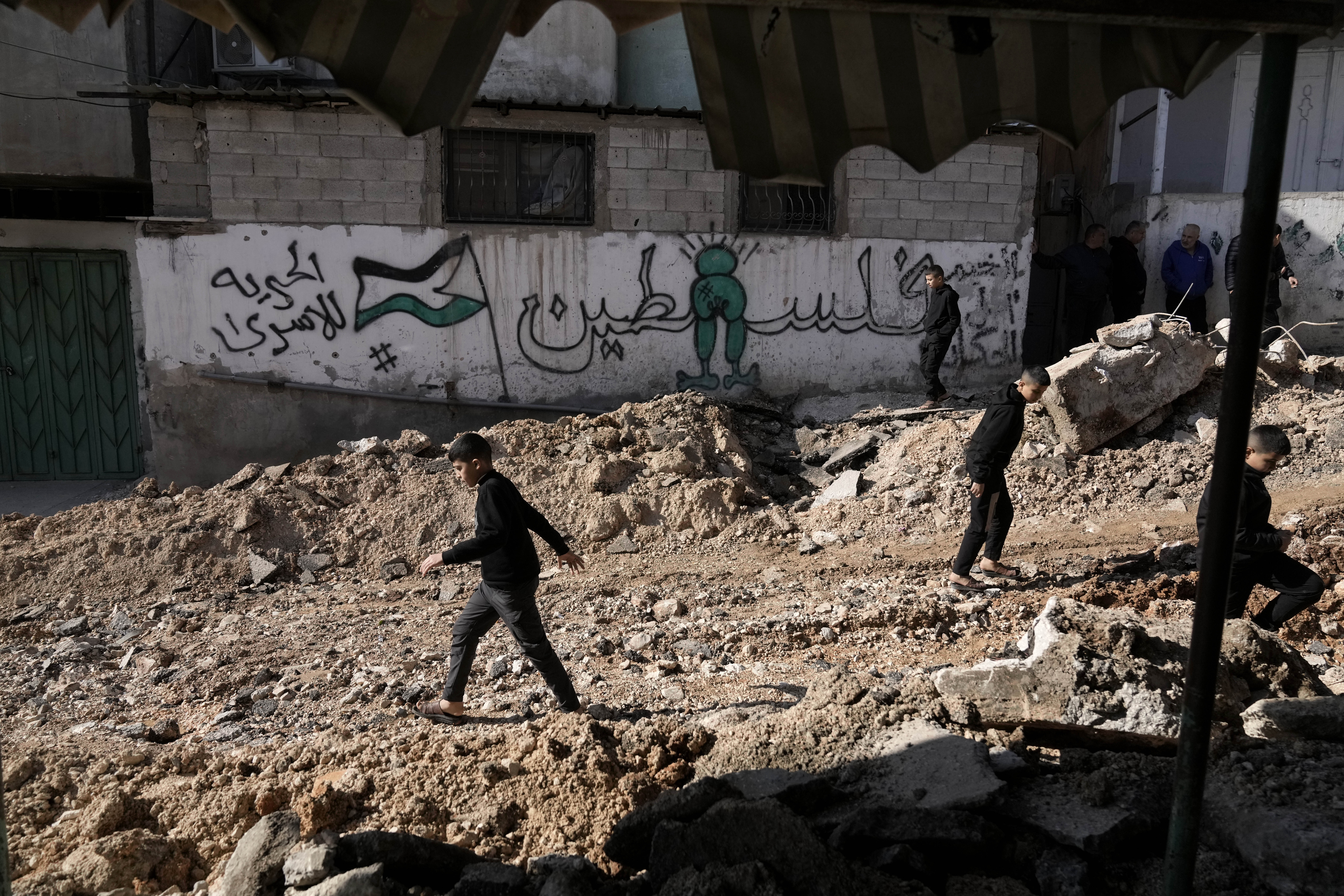 An Arabic mural reading, "Palestine," is the backdrop as Palestinian children walk on a street damaged in Israeli military operation at the Al-Fara'a refugee camp in the West Bank, Tuesday, Jan. 7, 2025. (AP Photo/Majdi Mohammed)