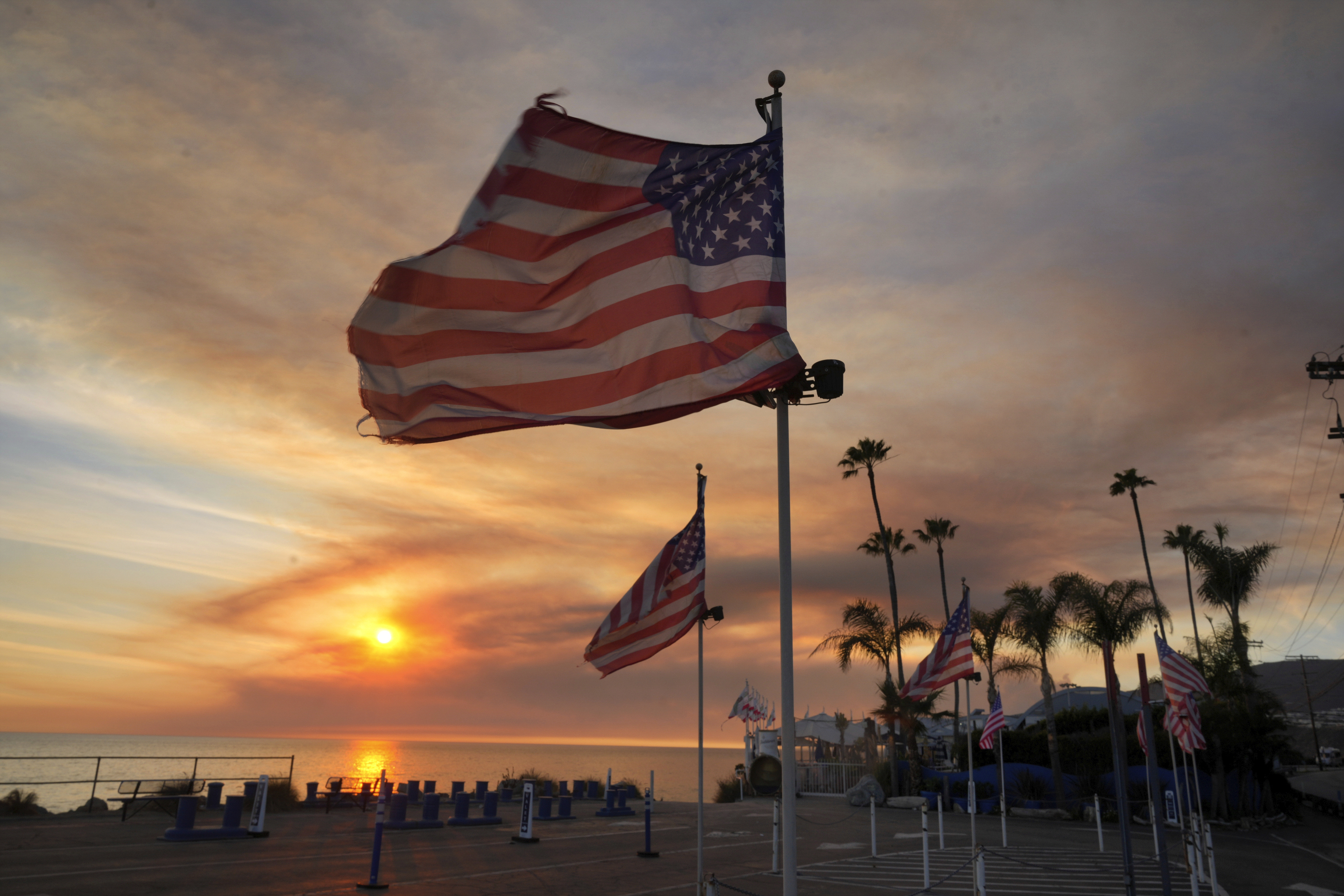 FILE - Flags fly under heavy winds before sunset as a plume of smoke from the Franklin Fire rises over the ocean Tuesday, Dec. 10, 2024, in Malibu, Calif. (AP Photo/Damian Dovarganes,File)