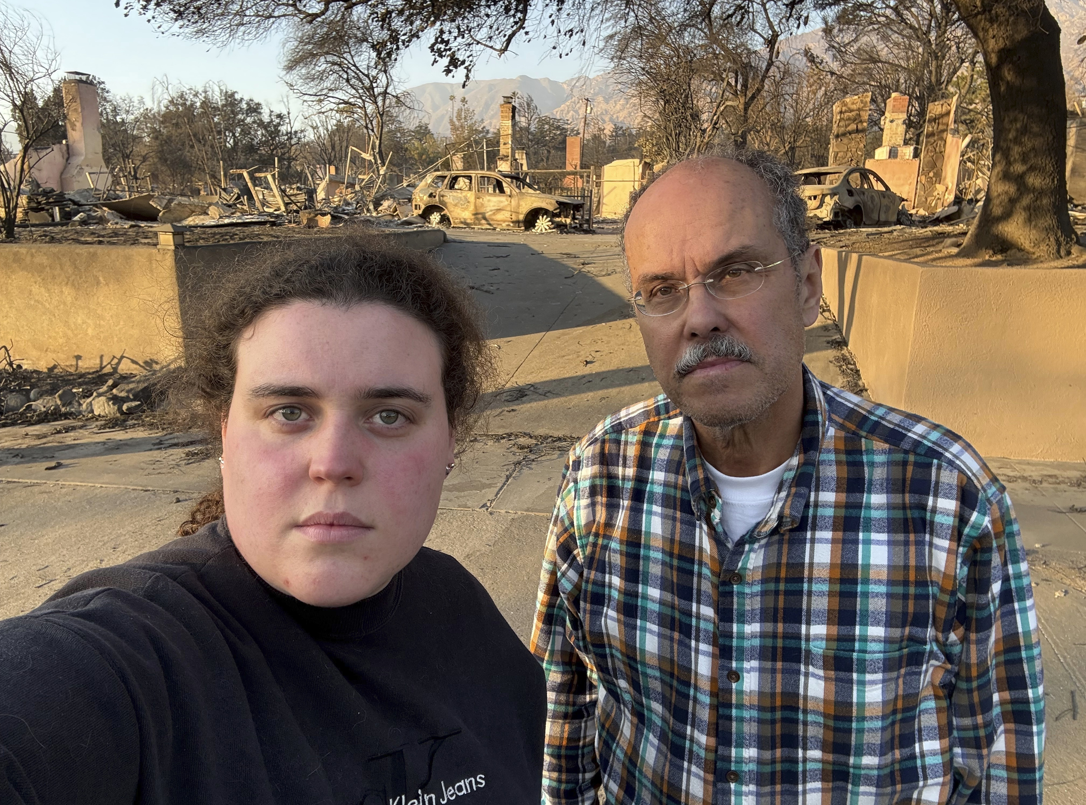 Vanessa Prata and her father, Aluizio Prata, pose for a self-portrait in Altadena, Calif., on Saturday, Jan. 11, 2025, with damage from the Eaton fire behind them. (Vanessa Prata via AP)